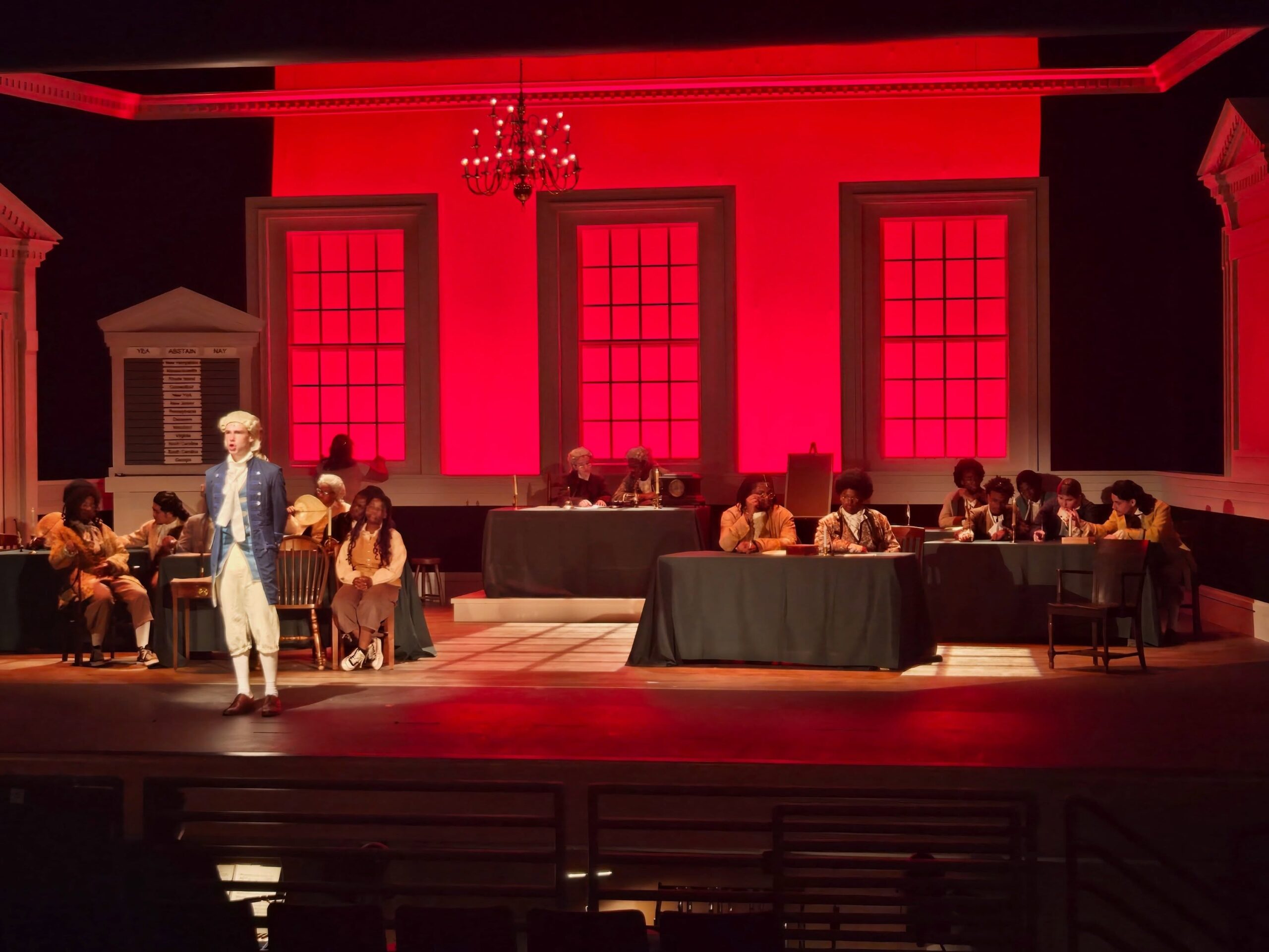Students perform a scene from 1776: The Musical on a stage designed as Independence Hall, with actors in colonial-era costumes gathered around tables beneath dramatic red lighting.