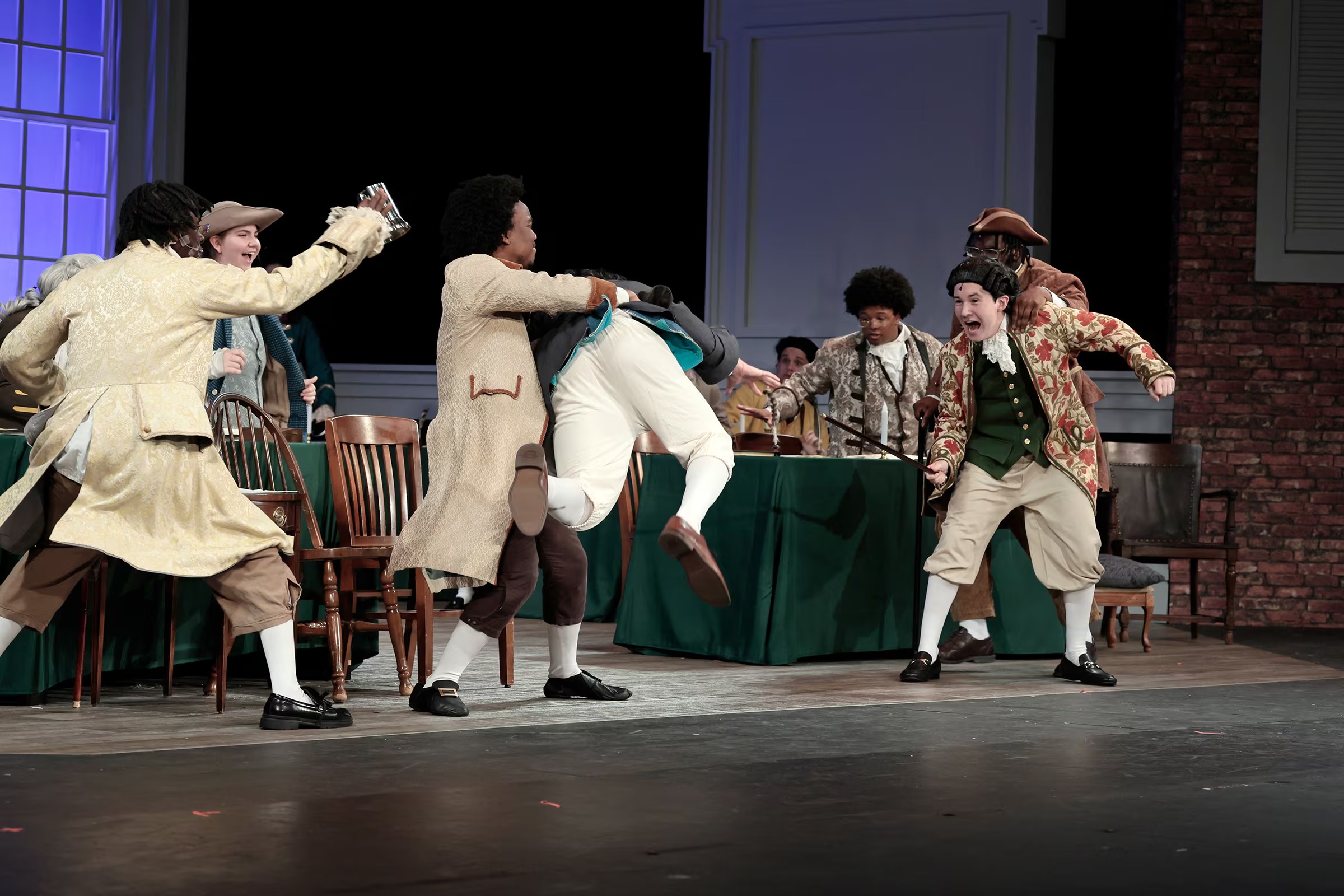 High school students in period costumes perform a lively scene from 1776: The Musical, with actors restraining one character while others react dramatically around a table onstage.