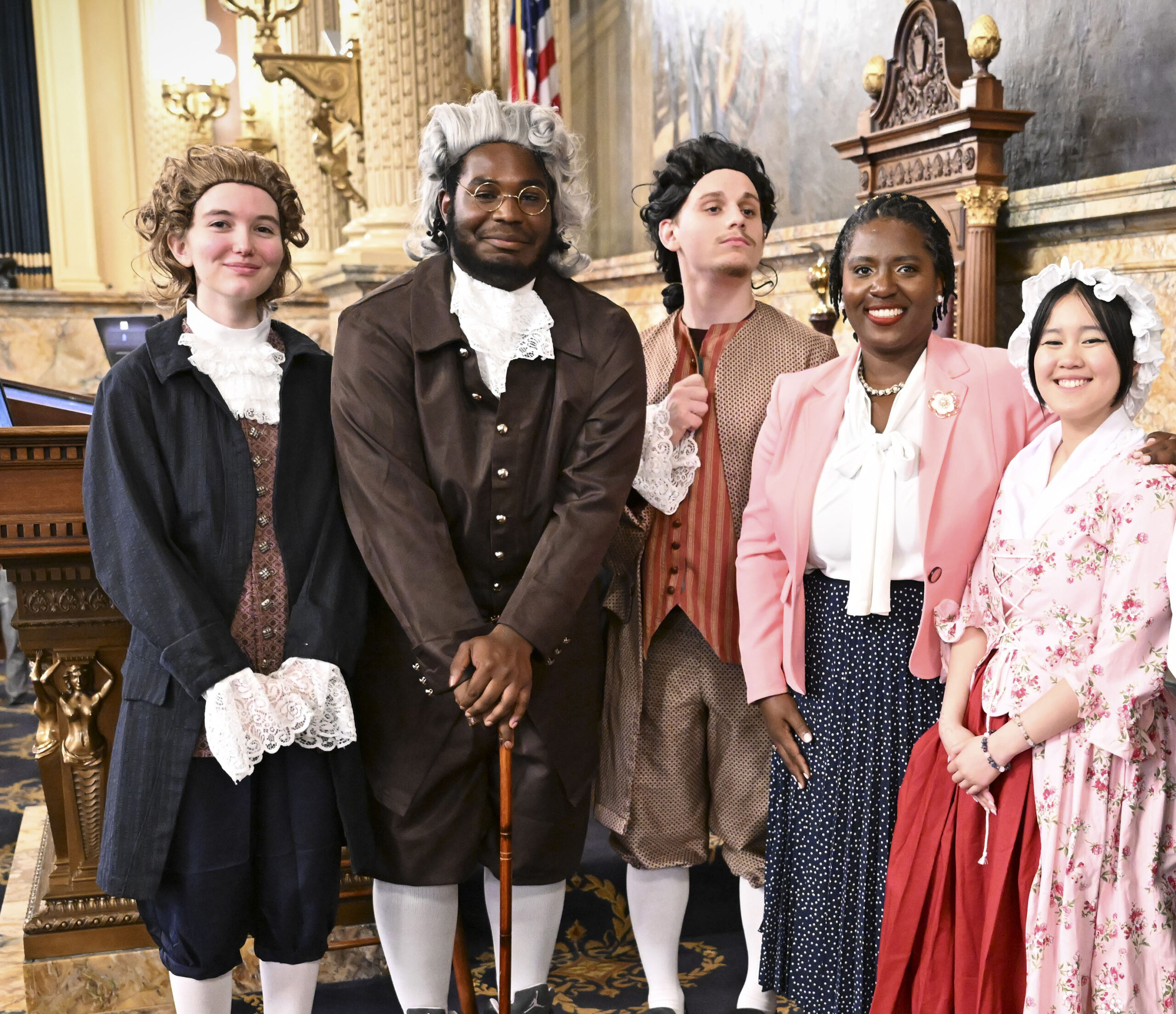 Students in colonial-era costumes pose with a woman in a pink blazer inside the Pennsylvania House chamber following a performance of 1776: The Musical.